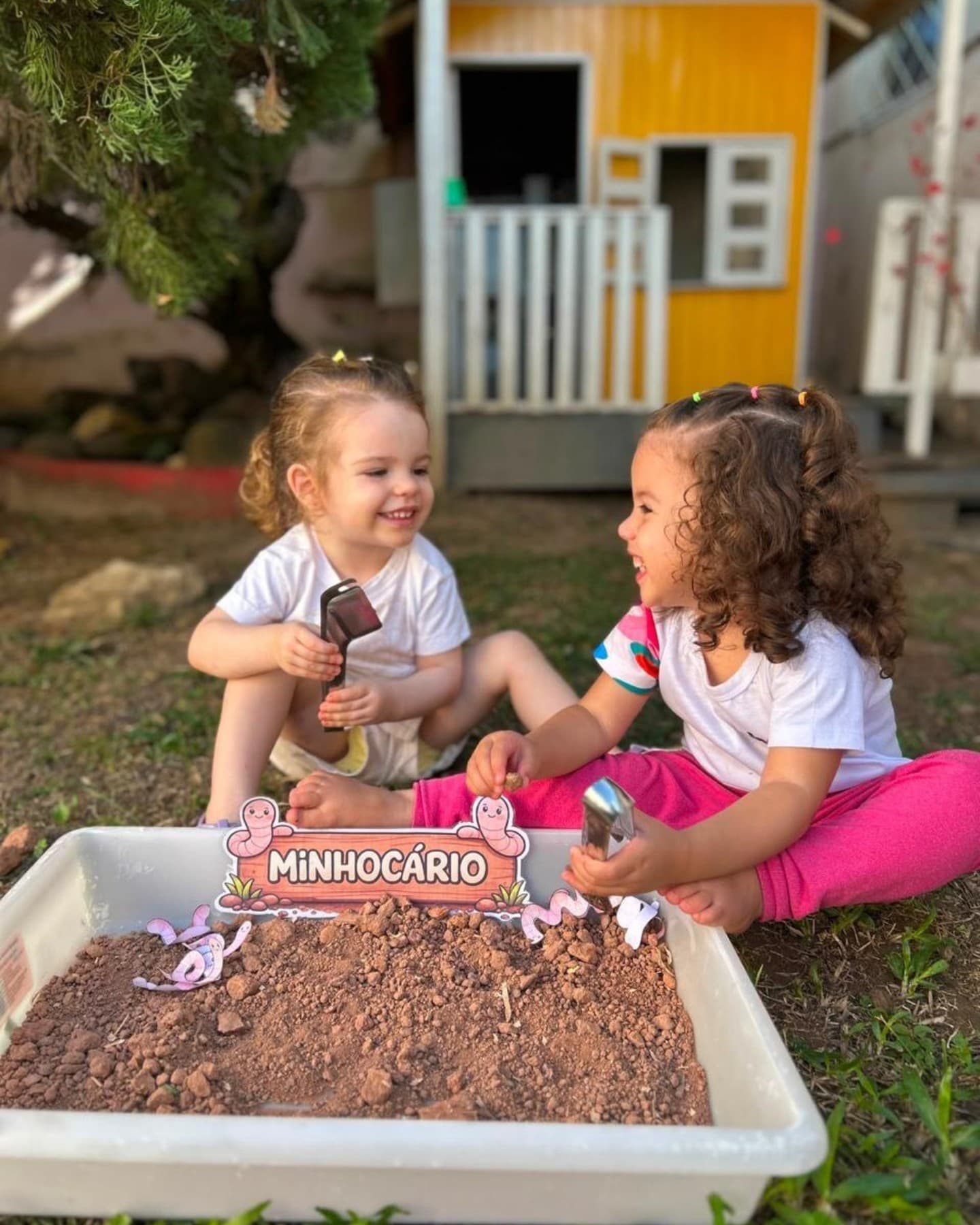 Duas meninas rindo sentadas na grama, com um minhocário de terra na frente